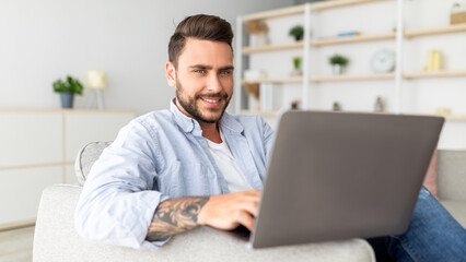 Fototapeta premium Positive guy reclining on sofa with laptop at home interior, panorama, copy space. Handsome man using notebook, watching movie, surfing internet or scrolling social networks