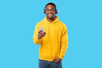 Cheerful Black Man Using Mobile Phone Wearing Earphones Listening To Music Online, Enjoying Playlist Posing Over Blue Studio Background, Smiling To Camera. Musical Application, People And Gadgets