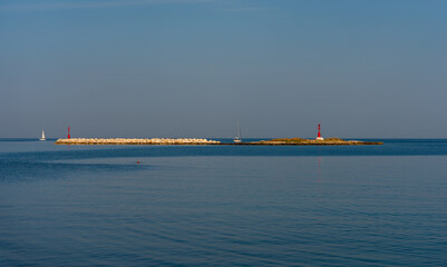 Porec, Croatia - August 13, 2025: Calm seascape with two distant red beacons on rocky breakwaters, sailboats on the horizon, and soft blue water under a clear sky.