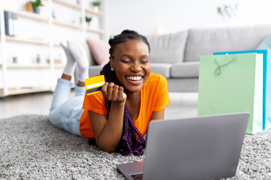 Young African American woman buying online with credit card and laptop, lying on floor at home. Millennial black lady doing remote shopping, using internet banking service, making contactless payment
