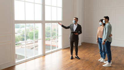 Male Realtor Or Architect Wearing Mask Showing View From New Empty House To Smiling Millennial Buyers, Pointing At Window. Arab Husband Hugging Wife, Visiting Residential Building Choosing Home