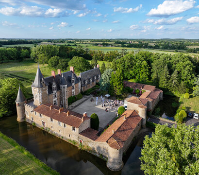 Aerial view of a medieval castle with its surrounding moat glinting under the sun, nestled among vibrant green trees and fields, Oree-d'Anjou, Pays de la Loire, France.
