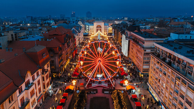 Aerial view of a dazzling Ferris wheel ablaze with light, standing at the heart of a festive Christmas market, with the Victory Square arch in the distance, Timisoara, Timis, Romania.