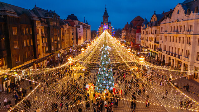 Aerial view of a dazzling Christmas tree and lights illuminating the bustling Victory Square, casting a warm glow on the Metropolitan Cathedral, Timisoara, Timis, Romania.