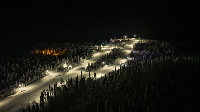 Aerial view of the snow-covered ski slopes under the embracing darkness, illuminated by the gentle glow of lights, Salla, Finland.