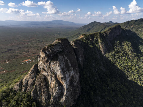 Aerial view of the rugged, rocky mountain ridge contrasting with the lush, green forest stretching towards the distant plains, Voi, Taita-Taveta County, Kenya.