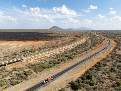Aerial view of a long, straight highway cutting through the arid landscape, with a train track running parallel, leading towards distant mountains, Voi, Taita-Taveta County, Kenya.