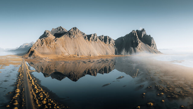 Aerial view of Vestrahorn Mountain's jagged peaks reflected in the tranquil lagoon, contrasting with the stark, winding road, Iceland.