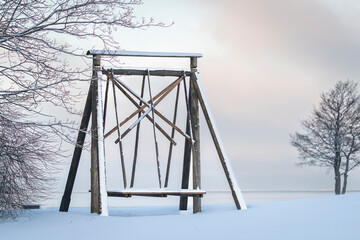 Minimalist winter scene with an empty wooden swing framed by bare trees and soft pastel sky. Snow-covered wooden swing standing alone in a quiet winter landscape by a frozen shoreline. 