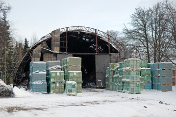 Burned industrial warehouse with stacked pallets wrapped in plastic, winter scene showing fire damage, storage and logistics aftermath.