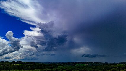The image shows a landscape of lush green hills and forests under a dramatic, cloud-laden sky, with areas of open land and what appears to be a column of rain in the distance.