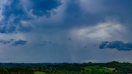 The image shows a landscape of lush green hills and forests under a dramatic, cloud-laden sky, with areas of open land and what appears to be a column of rain in the distance.