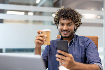Indian smiling young man wearing headphones sitting at a desk in the office, holding a cup of coffee and looking at the laptop screen