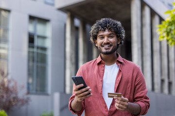 Portrait of a young smiling Indian man standing on the street, holding a phone and credit card and looking at the camera