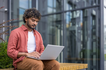 Serious young Indian man sitting on a bench outside, working and studying on a laptop