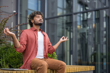 A young Indian man sits on a bench outside near a building with his eyes closed in a lotus position, resting relaxed