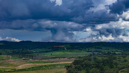 The image shows a landscape of lush green hills and forests under a dramatic, cloud-laden sky, with areas of open land and what appears to be a column of rain in the distance.