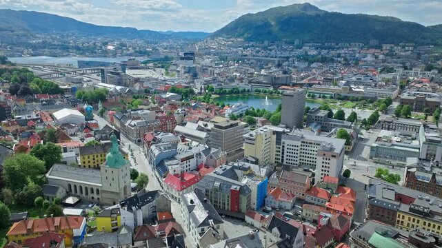 A wider aerial view of central Bergen, showcasing dense urban blocks, historic church towers, modern buildings, and the surrounding mountains fading into the distance under clear skies.