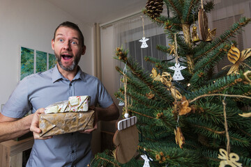 Man in shirt holding gift near charistmas tree. Shocked reaction on present. Holiday lifestyle background