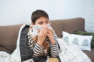 Sick child sitting on sofa wrapped in blanket, holding tissue to nose, winter illness concept, home interior, cold or flu symptoms.