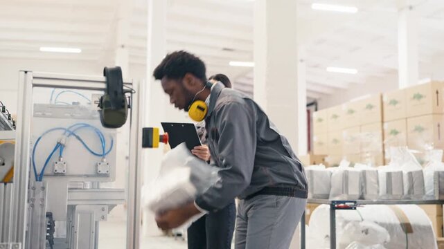 African American male factory worker wearing gray uniform and yellow headphones learning correct packaging procedure from female colleague supervising process beside machinery at warehouse.