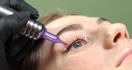 Beautician making permanent eyeliner makeup to young woman against green background, closeup
