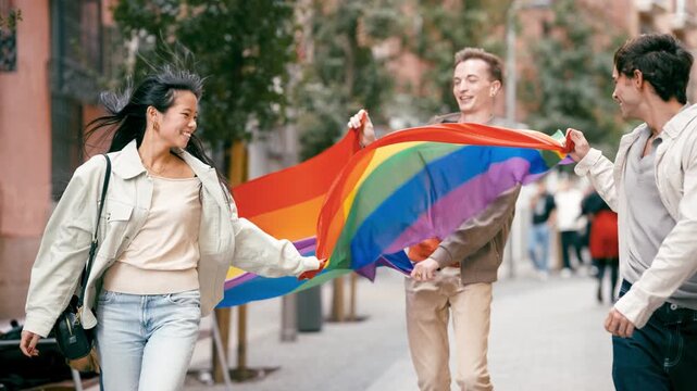 Diverse group of cheerful young friends smiling and walking together down a city street while proudly carrying a large, waving rainbow flag