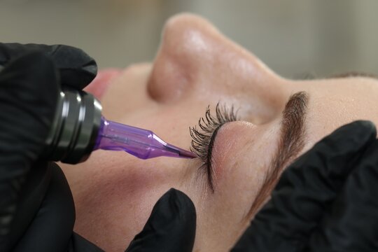 Beautician making permanent eyeliner makeup to young woman against blurred grey background, closeup