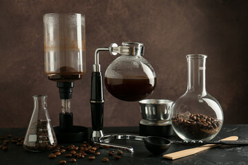 Siphon (vacuum) coffeemaker with coffee and glassware with beans on black table against brown background, closeup