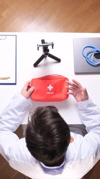 Vertical video medical professional in a white coat holding a red first aid kit, preparing to record an educational healthcare video with smartphone equipment on a white desk