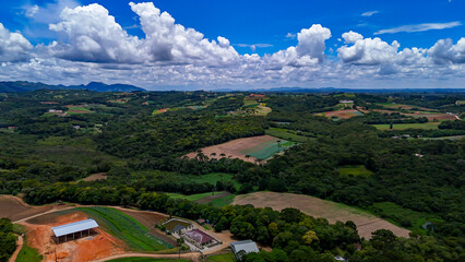 The image displays a vast rural and natural landscape with verdant hills, dense forest areas, cultivated fields, and a dramatic blue sky dotted with white and a few gray clouds.