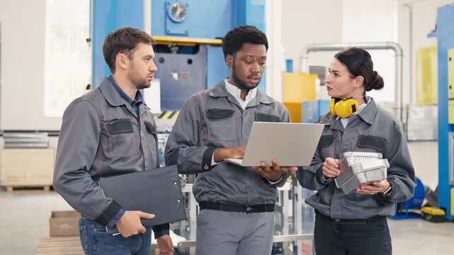 Male factory worker showing production data on laptop to colleagues. Woman holding foil containers checking quality. Man besides analyzing report. Team discussing manufacturing results in factory.