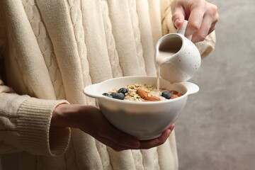 Woman pouring almond milk into bowl with oat flakes, nuts and blueberries on grey background,...
