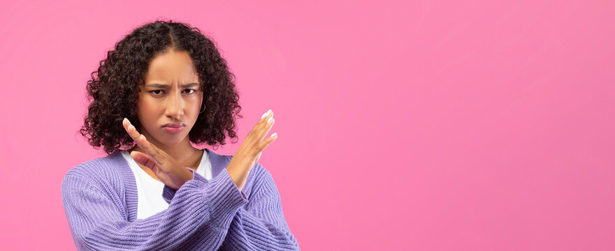 A woman stands with her arms crossed. She displays a serious expression and looks directly ahead. The background is a solid pink color, adding vibrancy to the setting.