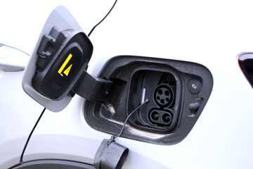 Open electric car charging port on white vehicle body, hinged plastic cover with warning symbol, black charging inlet with rubber seal, round connector and DC socket, modern eco automotive detail.