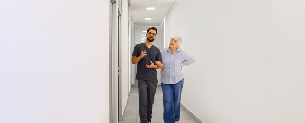 Male doctor walking arm in arm with senior woman explaining medical treatment while walking through hospital lobby