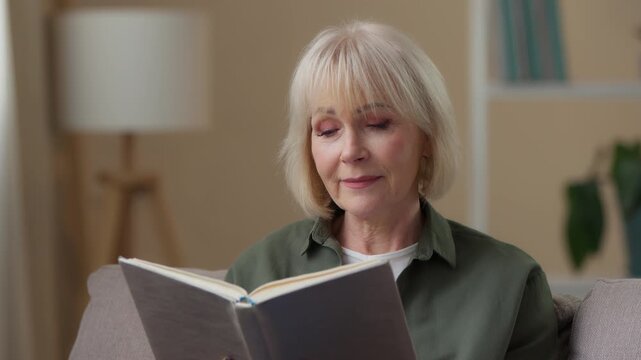 Calm peaceful Caucasian old woman reading book enjoying read literature at home looking away thinking mature lady senior female retired grandmother look on photo album memories remember on sofa indoor