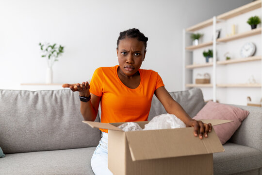 Wrong parcel. Confused black woman unpacking cardboard box, dissatisfied with bad delivery service, sitting on sofa at home. Young Afro lady unhappy about shipping mistake. Online shopping concept
