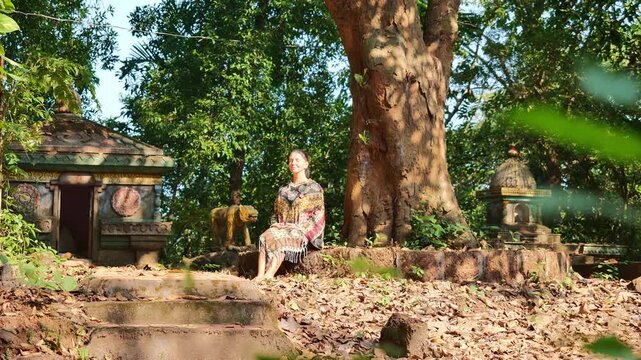 Middle-aged woman meditating in seated position under morning sunlight beside ancient shrine and large tree in dense forest, wearing colorful patterned shawl with eyes closed and hands in lap, express