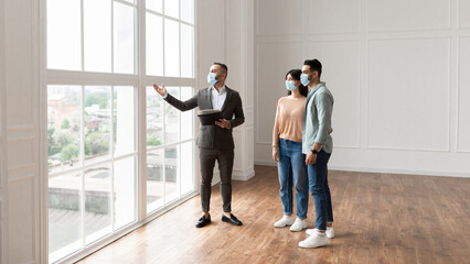 Sale. Real Estate Agent In Medical Mask Showing New Empty Apartment To Young Couple During Pandemic, Looking Pointing At Beautiful View From Big Panoramic Picture Windows. Happy Man And Woman Hugging