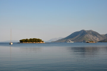 A lone sailboat floats on the calm Adriatic Sea near the pine covered Kosmac islet with mountains in the distance as seen from Vucine beach on Peljesac peninsula at dawn