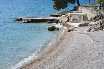 A scenic pebble beach in front of Emil Antica Street in Selce featuring a concrete sunbathing plateau and stone stairs leading to the clear blue Adriatic sea.