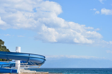 A large blue spiral water slide stands by the sea under a bright blue sky with white clouds on a sunny day in Selce.