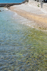 A scenic view of the pebble beach in front of Emil Antica Street in Selce featuring clear turquoise water and a stone seawall under bright sunlight.