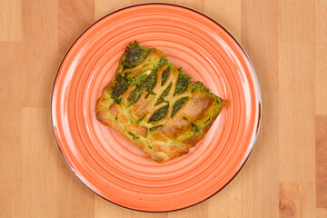 An overhead view of a slice of baked salmon and spinach in puff pastry, served on an orange plate on a wooden table