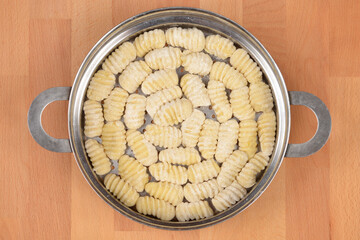 A top down view shows a full metal steamer basket of raw potato gnocchi dusted with flour and neatly arranged on a light wooden background.