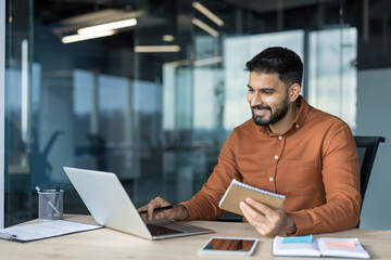 Young bearded indian male professional working on a laptop at a contemporary office desk, smiling while holding a notebook, and managing tasks with technology
