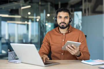 Young man wearing a headset, looking at the camera while working on a laptop and holding a tablet, managing remote customer support queries from a modern office environment