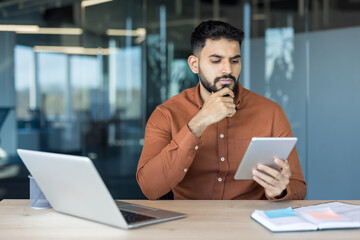 Young indian man in a modern office environment focused and thoughtfully reviewing information on a digital tablet, demonstrating concentration and problem-solving skills at work