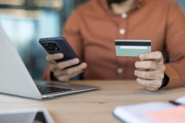 Person holding credit card and smartphone while completing secure online purchase or banking on a laptop at a wooden desk, close-up hands, cashless convenience and privacy-focused transaction